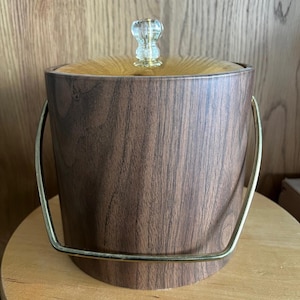 May include: A vintage ice bucket with a faux wood grain finish. The bucket has a gold-colored handle and a clear, decorative knob on the lid. The interior of the lid is a light amber color.