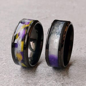 Può includere: Due anelli neri con intarsi colorati. Uno presenta un mix di giallo, viola e bianco, mentre l'altro ha un design bianco, grigio e viola. Gli anelli sono su uno sfondo grigio.