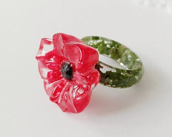 May include: A ring featuring a vibrant red poppy flower with a dark center, attached to a band that has a textured, mossy green appearance. The flower petals are translucent and layered, creating a realistic floral look on the finger.
