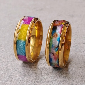 May include: Two gold-toned metal rings featuring a wide band design. The left ring has a central inlay of segmented bright pink, yellow, and blue resin. The right ring features a central inlay of multicoloured, marbled resin with swirls of blue, pink, and gold.