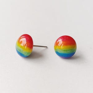 May include: Two rainbow-colored stud earrings. Each earring is a small, rounded shape with a post. The colors of the rainbow are visible, transitioning from red to orange, yellow, green, blue, and purple. The earrings are set against a white background.