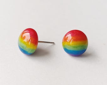 Può includere: Due orecchini a bottone arcobaleno. Ogni orecchino è di forma piccola e arrotondata con un perno. I colori dell'arcobaleno sono visibili, passando dal rosso all'arancione, giallo, verde, blu e viola. Su sfondo bianco.