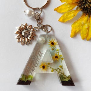 May include: A personalized keychain featuring the letter "A" with embedded yellow sunflowers and the name "Anika". The keychain includes a silver sunflower charm, a pearl bead, and a silver keyring. The base of the letter is dark brown.