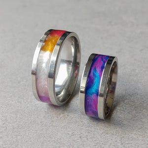 Può includere: Due anelli in metallo con fasce color argento lucido. L'anello più grande presenta un intarsio in resina con motivi vorticosi nei toni del rosa, arancione e bianco. L'anello più piccolo presenta un intarsio in resina nei toni del blu e viola.