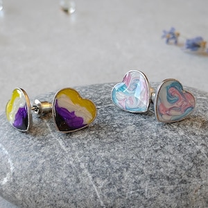 May include: Two pairs of heart-shaped stud earrings. One pair features a non-binary pride flag design with yellow, white, purple, and black colors. The other pair has a swirl of pink and blue. The earrings have a silver border and are displayed on a gray stone.