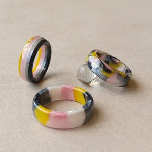 May include: Three circular rings featuring a smooth, polished finish with a marbled colour pattern of yellow, pink, white, grey, and black. One ring stands upright, while the other two lie flat, accompanied by a small, clear, spherical glass bead.