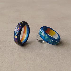 May include: Two rings with smooth, rounded bands. The left ring is dark navy blue with gold star accents and a rainbow-coloured interior. The right ring is a lighter blue with white speckles and a rainbow-coloured interior, resting on a clear sphere.