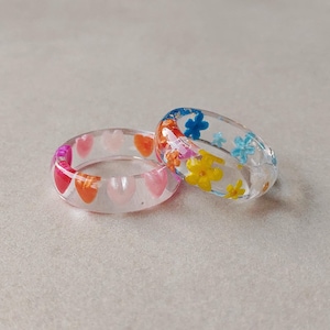 May include: Two clear resin rings. One ring features embedded small pink, red, and orange heart shapes. The second ring contains embedded colorful miniature flower shapes in shades of yellow, blue, orange, and pink throughout the band.