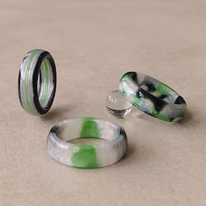 Op de afbeelding: Drie ringen met een gemarmerd patroon van zwart, wit en felgroen. Eén ring staat rechtop, één ligt plat en één staat schuin, vergezeld door een kleine, doorzichtige, bolvormige kraal. De ringen hebben een gladde, gepolijste afwerking.
