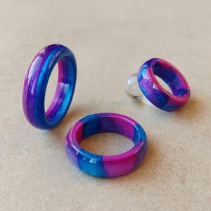 Puede incluir: Tres anillos de resina en tonos azules, morados y rosas. Los anillos tienen un efecto veteado, con los colores arremolinándose. Los anillos se muestran sobre una superficie neutra.