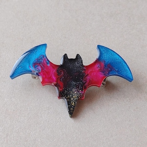 May include: A bat-shaped brooch featuring a glossy resin finish with a swirling color gradient. The center is black with gold glitter, transitioning into vibrant magenta and bright blue at the wingtips. The surface has a smooth, reflective texture.