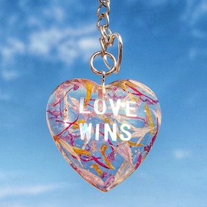 Könnte beinhalten: Herzförmiger Schlüsselanhänger mit den Worten "LOVE WINS" in klarem Harz eingebettet. Der Schlüsselanhänger ist mit bunten Trockenblumen gefüllt und hat eine silberfarbene Metallkette und einen Schlüsselring. Der Hintergrund ist ein strahlend blauer Himmel.