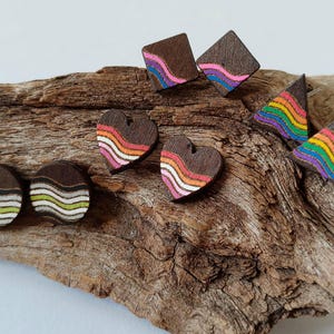 May include: A collection of wooden earrings in various shapes, including circles, hearts, squares, and triangles. Each earring features a dark brown base with colorful, wavy stripes in shades of white, pink, orange, yellow, green, blue, and purple. The earrings are displayed on a piece of driftwood.