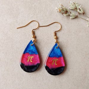 Puede incluir: Un par de pendientes en forma de lágrima con ganchos dorados. Los pendientes presentan un degradado de color azul, rosa y negro, con un símbolo pi dorado en el centro. Se muestran sobre un fondo neutro.