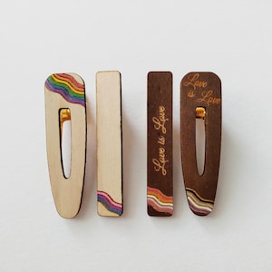 Puede incluir: Cuatro horquillas de madera con cierres metálicos. Dos horquillas son de madera clara, una con un diseño de onda arcoíris. Dos horquillas son de madera oscura, una con un diseño de onda arcoíris y otra con el texto grabado Love is Love.