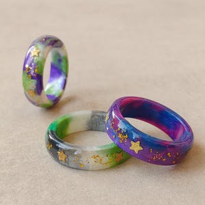 Op de afbeelding: Drie harsringen met wervelende patronen in paars, blauw, groen en wit. Elke ring is ingelegd met kleine gouden stervormige confetti en fijne gouden glitterdeeltjes, wat een hemels effect creëert over de gladde, afgeronde ringbanden.