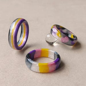 Op de afbeelding: Drie ringen gemaakt van gladde, gepolijste hars. Eén ring staat rechtop, terwijl twee ringen plat liggen. De ringen hebben horizontale strepen in paars, wit, geel en zwart, wat zorgt voor een levendige, veelkleurige uitstraling met een glanzende afwerking.