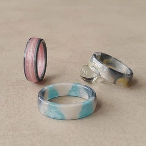 Op de afbeelding: Drie harsringen worden getoond. Eén ring staat rechtop met verticale roze en grijze strepen. Een tweede ring ligt plat en heeft een wit en lichtblauw gemarmerd patroon. De derde ring staat schuin en toont grijze, witte en gele wervelingen.