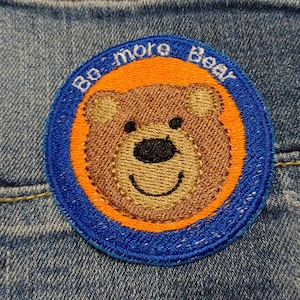Pode incluir: Um remendo bordado azul e laranja com um rosto de urso marrom e o texto "Be More Bear".