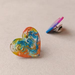 May include: A heart-shaped pin featuring a marbled swirl of orange, blue, yellow, and white colors with embedded gold glitter, a small gold crescent moon, and a gold star charm. A second pin with a pink and blue gradient is visible in the background.