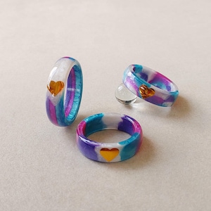 Pode incluir: Três anéis com um padrão de redemoinho nas cores branco, roxo e azul. Cada anel tem um acabamento liso e polido e é acentuado com um pequeno coração de cor dourada metálica incrustado na superfície externa da banda do anel.
