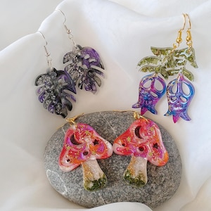 以下が含まれることがあります： 異なるデザインのイヤリングが3ペア。最初のペアは、紫と黒の葉のデザイン。2番目のペアは、緑と紫の花のデザイン。3番目のペアは、赤と白のキノコのデザインです。