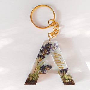 May include: A gold-coloured keyring featuring a clear resin letter "A" with dried lavender and a small butterfly. The name "Aida" is written in gold script on the resin. The keyring has a gold ring and chain.