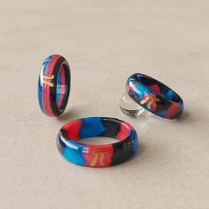 Op de afbeelding: Drie ringen van hars met een wervelend patroon van blauwe, rode en zwarte kleuren. Elke ring heeft een goudkleurig pi-symbool ingebed in het buitenoppervlak. De ringen zijn gerangschikt met één rechtopstaand, één platliggend en één schuin.