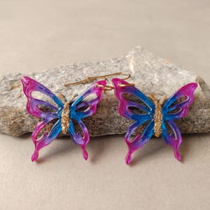 Peut inclure: Une paire de boucles d'oreilles en forme de papillon présentant un dégradé translucide de couleurs rose et bleu vives. Le centre de chaque papillon est détaillé avec une finition dorée texturée, et chaque boucle inclut un crochet en métal doré.