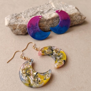 May include: Two pairs of crescent moon earrings. One pair features a gradient of magenta, purple, and blue resin. The second pair is made of clear resin with yellow, black, and white swirls, accented with small gold star-shaped glitter flakes.