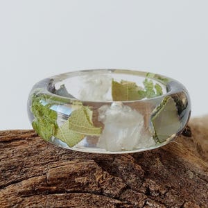 Puede incluir: Un anillo de resina transparente con hojas verdes y cuarzo blanco incrustados. El anillo está sobre un trozo de madera flotante.