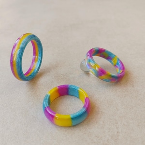Könnte beinhalten: Drei Ringe aus glattem, poliertem Material mit einer lebendigen Farbpalette aus leuchtendem Pink, sonnigem Gelb und Himmelblau. Die Ringe weisen ein marmoriertes oder gewirbeltes Muster auf ihren kreisförmigen Bändern auf und glänzen.