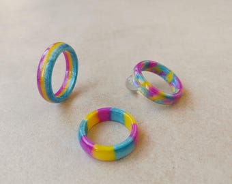 Puede incluir: Tres anillos hechos de material liso y pulido con una paleta de colores vibrantes de rosa brillante, amarillo soleado y azul cielo. Los anillos muestran un patrón jaspeado o arremolinado en sus bandas circulares con un acabado brillante.
