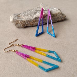 Pode incluir: Dois pares de brincos pendentes com armações triangulares abertas. Um par apresenta um degradê de rosa para azul, enquanto o outro exibe um degradê tricolor de rosa, amarelo e azul. Ambos os conjuntos incluem ganchos de metal dourado.