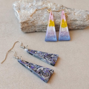 Könnte beinhalten: Zwei Paar dreieckige Ohrhänger mit Metallhaken. Ein Paar zeigt einen Farbverlauf aus Pink, Gelb, Weiß und Blau. Das zweite Paar hat ein dunkles, marmoriertes Muster in Schwarz, Violett und Silber mit kleinen, eingebetteten sternförmigen Akzenten.