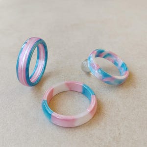 Peut inclure: Trois bagues circulaires avec une finition lisse et polie. Une bague présente des rayures verticales roses, blanches et bleues. Les deux autres bagues présentent un motif marbré et tourbillonnant de couleurs rose, blanche et bleue.