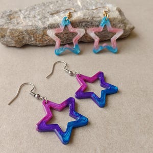 Puede incluir: Dos pares de pendientes colgantes con forma de estrella. Un par presenta un diseño de resina veteada en rosa, blanco y azul con ganchos dorados. El segundo par presenta un diseño de resina veteada en morado, magenta y azul con ganchos plateados.