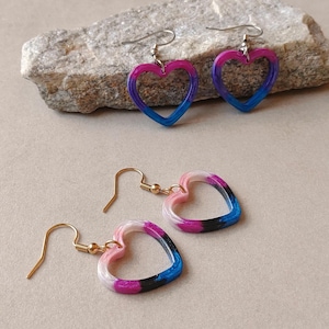 May include: Two pairs of heart-shaped dangle earrings. One pair features a gradient of purple and blue, while the other pair displays a blend of white, pink, purple, black, and blue. Each earring hangs from a simple metal hook wire.