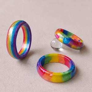 May include: Three rings featuring vibrant rainbow color patterns. One ring stands upright displaying vertical stripes of red, orange, yellow, green, blue, and purple. Two other rings lie flat, showcasing a multi-colored, blended, and swirled design.
