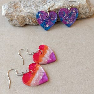 Pode incluir: Dois pares de brincos em forma de coração. Um par apresenta um padrão de redemoinhos azuis e roxos com glitter prateado em forma de estrela. O segundo par exibe um gradiente de redemoinhos vermelhos, laranja, brancos e rosas com acabamento liso.