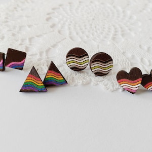 Può includere: Orecchini a bottone in legno di varie forme: quadrati, triangoli, cerchi e cuori. Ogni orecchino è marrone scuro con disegni colorati dipinti. Gli orecchini quadrati hanno un'onda rosa e viola. I triangoli hanno strisce arcobaleno. I cerchi hanno onde verdi e bianche. I cuori hanno strisce arancioni e rosa.