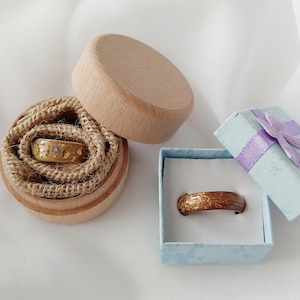 May include: Two rings, one in a wooden box with a jute lining and the other in a light blue gift box with a purple ribbon. The rings have gold and brown tones. One ring has a small gem.