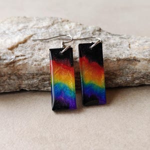 May include: Two rectangular earrings with a rainbow gradient design. The earrings feature a black top and a vibrant blend of colours including red, orange, yellow, green, blue, and purple. They hang from silver-coloured ear wires.