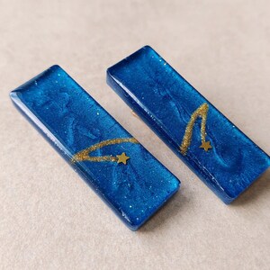 May include: Two rectangular, blue resin barrettes with gold glitter and a gold star and curved line design. The barrettes have a glossy finish and are set against a neutral background.