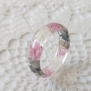 May include: A clear resin ring featuring pink and grey floral accents. The ring is made with real pressed flowers and is a unique piece of jewellery.