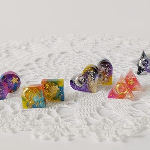 May include: Assortment of colorful resin earrings with celestial and geometric designs. The earrings feature a variety of shapes, including hearts, squares, triangles, and stars. The resin is translucent and contains glitter and gold accents.