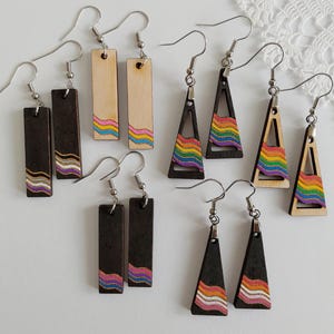 Pode incluir: Conjunto de oito brincos de madeira com listras arco-íris. Os brincos são em forma de retângulos e triângulos. Alguns são pretos e outros são de madeira clara.