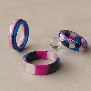 Peut inclure: Trois bagues en résine présentant un motif marbré dans des tons de rose, bleu, noir et blanc. Une bague est debout, une est posée à plat et une est inclinée, accompagnées d'une petite perle de verre sphérique et transparente entre elles.