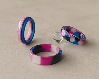 Puede incluir: Tres anillos de resina con una paleta de colores rosa, blanco, azul y negro. Un anillo está en posición vertical, otro plano y el tercero inclinado, mostrando un patrón jaspeado o de rayas. Una pequeña cuenta esférica transparente está cerca.