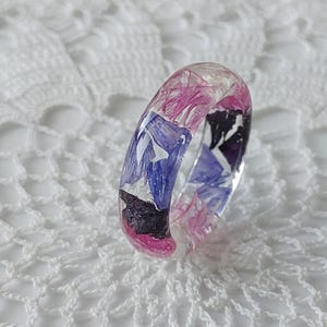 以下が含まれることがあります： ピンク、パープル、ブルーのドライフラワーが埋め込まれた透明なレジンリングです。